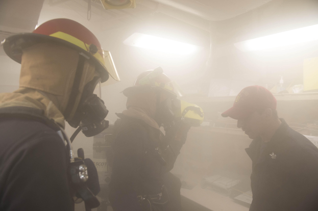 Feuerwehrleute in einem raucherfüllten Raum während einer Übung, einer hält eine Kamera, trägt Helme mit sichtbaren Lichtern im Hintergrund.