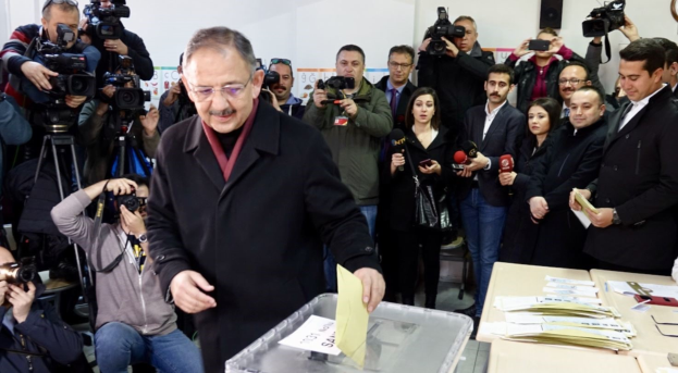 Ein Mann in einem schwarzen Mantel und Brille stimmt an einer Wahlurne ab, umgeben von Menschen mit Kameras und Mikrofonen, Tischen mit Papieren und Gegenständen und einer Wand mit einer Uhr und einem Lautsprecher im Hintergrund.