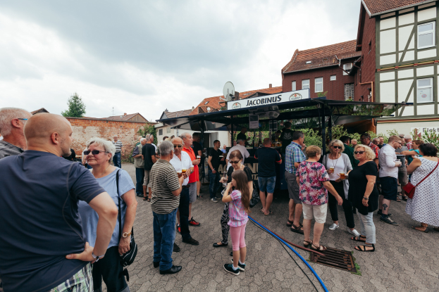 Gruppe von Menschen auf einem Outdoor-Bierfestival vor einem Gebäude mit Fenstern, Bäumen und einem Schuppen mit einem Schild im Hintergrund.