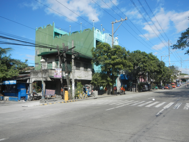 Eine Stadtstraße mit einem grünen Gebäude, fahrende Fahrzeuge, Fußgänger auf dem Gehweg und Strommasten mit Drähten unter einem bewölkten Himmel.