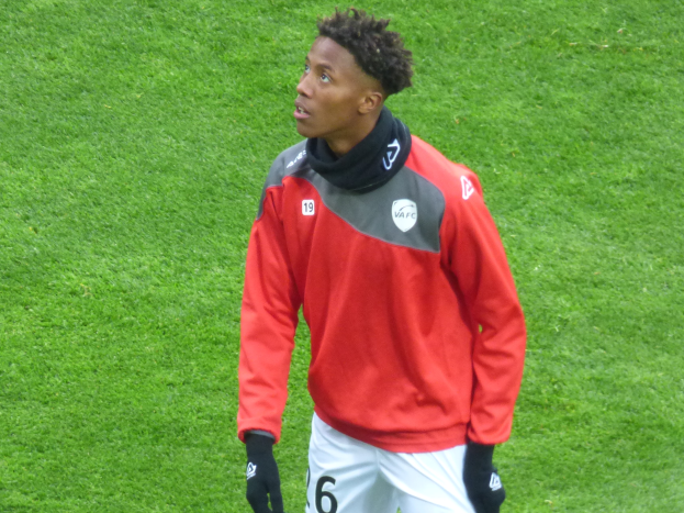 Ein junger Mann in einer roten Jacke und weißen Shorts steht auf einem Fußballfeld und schaut rechts mit entschlossener Miene, Handschuhe tragend.