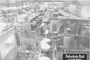 Schwarzes und weiße Foto eines großeen Fabrikinneren mit einer Person, die zwischen Industrieanlagen, Rohren und konstruktiven Elementen steht, beschriftet mit "Arbeidets Rett, die größteste Fabrik der Welt."