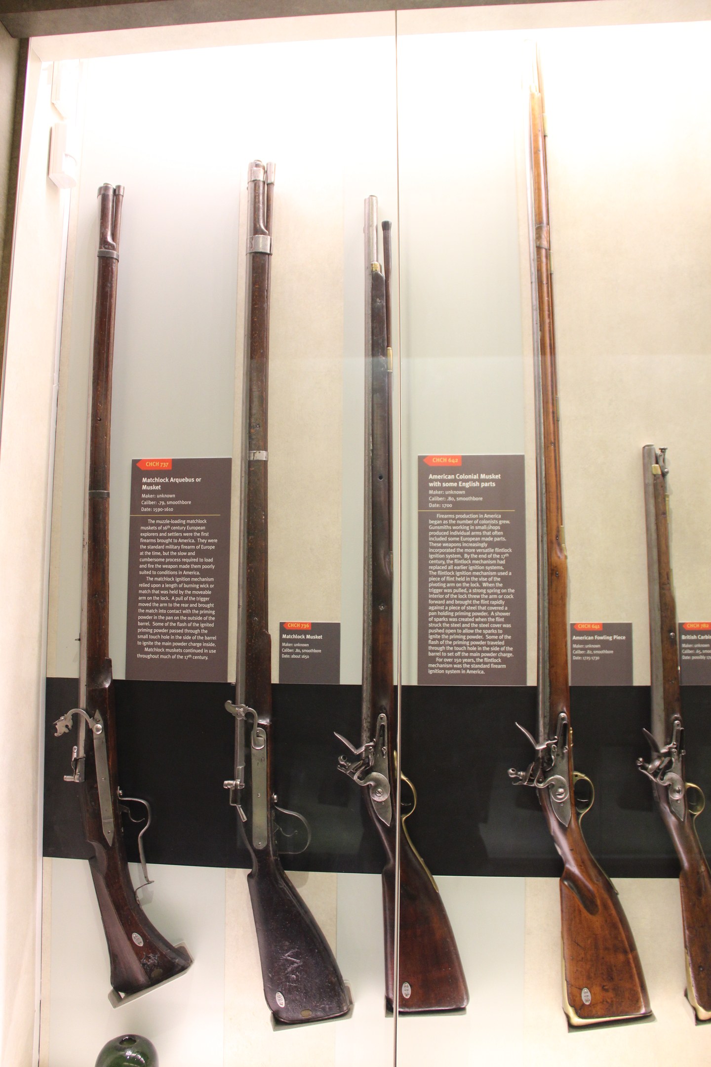Vitrine mit verschiedenen Feuerwaffen, darunter ein Steinschlossgewehr, in einer Reihe mit Informationsschildern dahinter und einem unbestimmten Objekt unten.