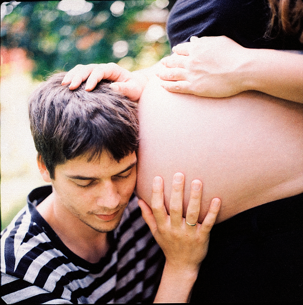 Ein Mann und eine Frau stehen nebeneinander, der Mann berührt sanft den Bauch der Frau und schaut auf ihr ungeborenes Kind.