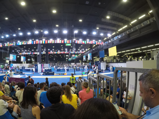 Eine große Menschenmenge sitzt und steht in einer Indoor-Sportarena, mit Metallzaun auf der rechten Seite, Bildschirmen, Flaggen und Deckenleuchten im Hintergrund.