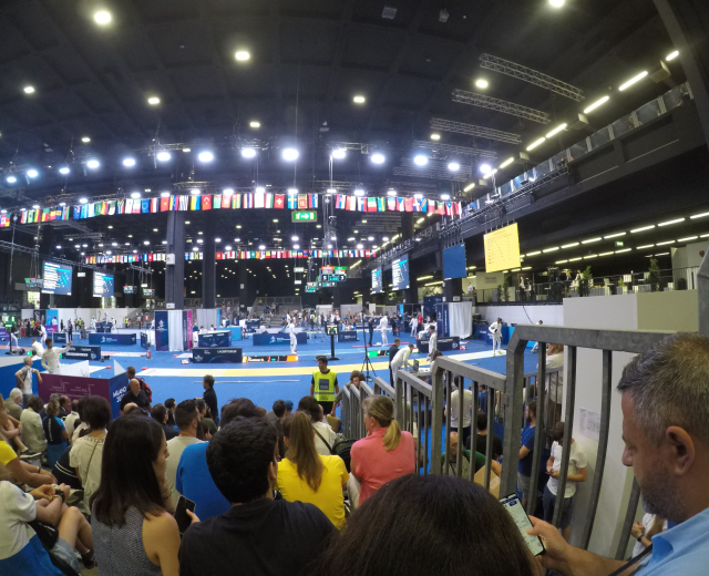 Eine große Menschenmenge sitzt und steht in einer Indoor-Sportarena, mit Metallzaun auf der rechten Seite, Bildschirmen, Flaggen und Deckenleuchten im Hintergrund.