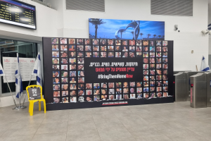 Große Wand mit mehreren Fotos, gelber Stuhl, israelische Regierungsflaggen auf Ständern, eine Tafel mit dem Text "Bring Them Home Now", ein Wandbildschirm, Deckenleuchten und andere Gegenstände.