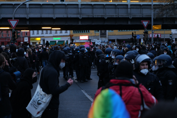Eine große Gruppe von Menschen vor einer Reihe von Polizisten, einige mit Helmen und Taschen oder Kameras, mit Gebäuden, Schildern, Pfosten, Verkehrszeichen, einer Brücke und einem Baum im Hintergrund.