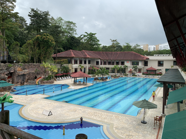 Großes Schwimmbad im Ritz-Carlton, Kota Kinabalu, mit Menschen, Sonnenschirmen, Stühlen, Tischen, Pflanzen, Bäumen, Felsen, Gebäuden, einem Schuppen auf der rechten Seite und einem sichtbaren Himmel im Hintergrund.