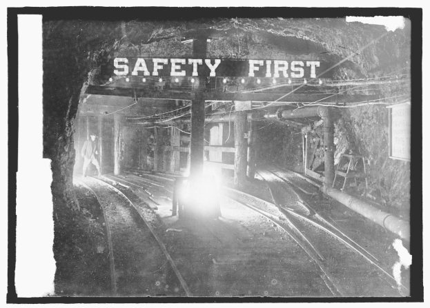 Schwarzes und weißes Foto eines Zuges, der durch einen Tunnel fährt, mit einer Person auf dem Gleis im Vordergrund, Masten und Lichter im Hintergrund und einem "Sicherheit geht vor"-Schild.
