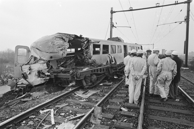 Schwarz-weißes Bild einer Zugkollision auf den Schienen mit Einsatzkräften in Helmen und einem Hintergrund aus Strommasten, Bäumen, Gebäuden und einem klaren Himmel.