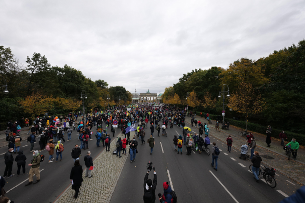 Eine große Gruppe von Menschen marschiert auf einer von Bäumen gesäumten Straße mit Laternenmasten, hält Kameras, während einer Protestdemo in Berlin mit einem Gebäude und einem klaren Himmel im Hintergrund.