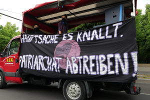 Roter Lkw mit einer Fahne, die eine Straße mit Bäumen entlangfährt, unter einem klaren blauen Himmel, zwei maskierte Personen sind im Inneren sichtbar.