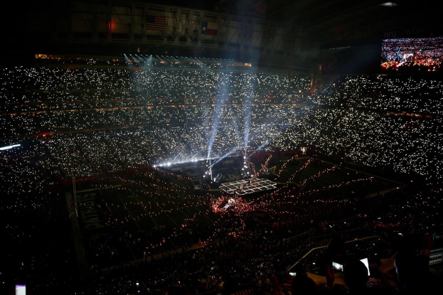 Große Menschenmenge in einem hell erleuchteten Stadion bei Nacht, mit einem Bildschirm auf der rechten Seite, der die Ergebnisse der Super Bowl XLVII-Halbzeitshow anzeigt.