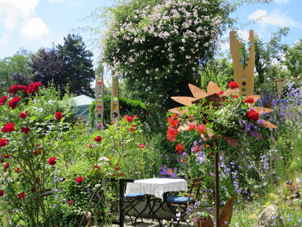 Ein Garten mit bunten Blumen und Pflanzen, mit einem Tisch und Stühlen mit einer Decke, vor Bäumen und einem bewölkten Himmel auf dem Chelsea Flower Show.