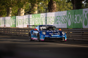 Blauer Porsche 911 GTS auf einer Rennstrecke mit Absperrung und Bannern, Bäumen und einem klaren Himmel im Hintergrund.