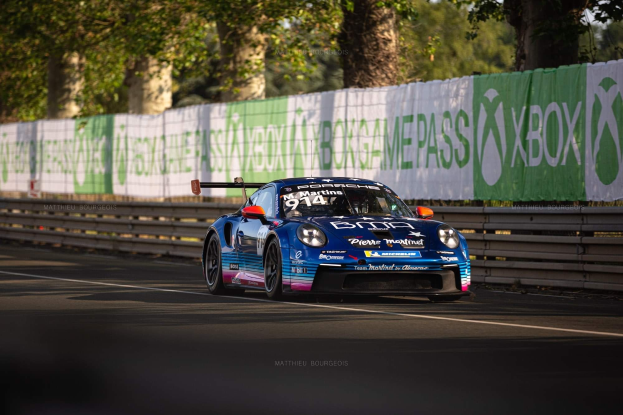 Blauer Porsche 911 GTS auf einer Rennstrecke mit Absperrung und Bannern, Bäumen und einem klaren Himmel im Hintergrund.
