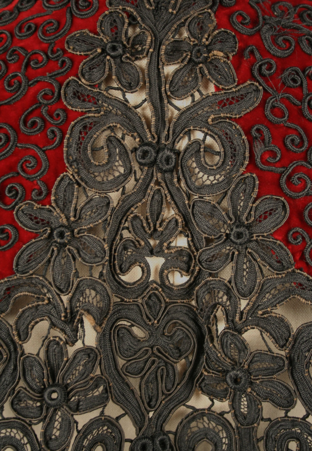 Nahaufnahme eines roten Kleids mit schwarzer Litzen-Überlagerung, das detaillierte Litzen-Muster auf rotem Stoff zeigt.