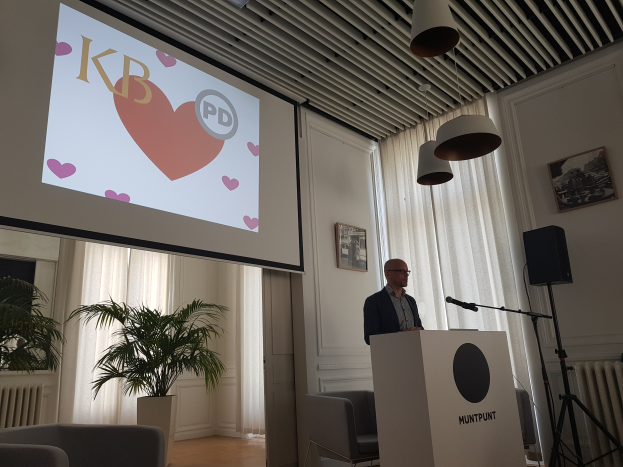 Ein Mann steht an einem Podium mit einem Mikrofon und hält eine Präsentation vor einem Projektionsschirm, mit Stühlen, einer Pflanze, Fotorahmen, Vorhängen und Deckenlampen im Raum.