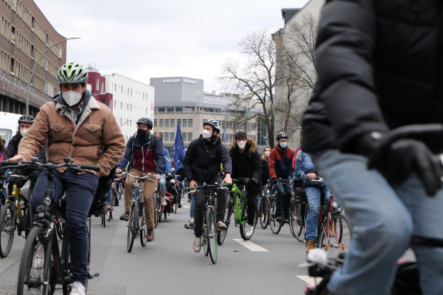 Gruppe von Radfahrern in Helmen und Handschuhen auf einer von Bäumen gesäumten Straße in Berlin unterwegs, mit Gebäuden und einem geparkten Fahrzeug im Hintergrund.