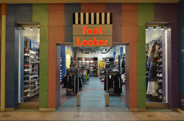Innenansicht eines Foot Locker Schuhgeschäfts mit Kleidung an Bügeln, Waren auf Gestellen und Deckenbeleuchtung, die den Boden zeigt.