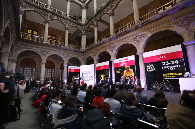 Eine Gruppe von Menschen, die an einem Tisch mit einem Podium und einem Mikrofon vor einer Menge, Kameras und Bannern sitzen, was eine Pressekonferenz für das CDMX 2017-Ereignis vermuten lässt.