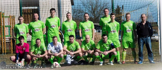 Eine Gruppe von Männern in grünen T-Shirts steht auf einem Fußballfeld, einige sind in der Hocke, mit einem Ball und einem Torpfosten; der Hintergrund enthält Bäume, Gebäude und Fahrzeuge.