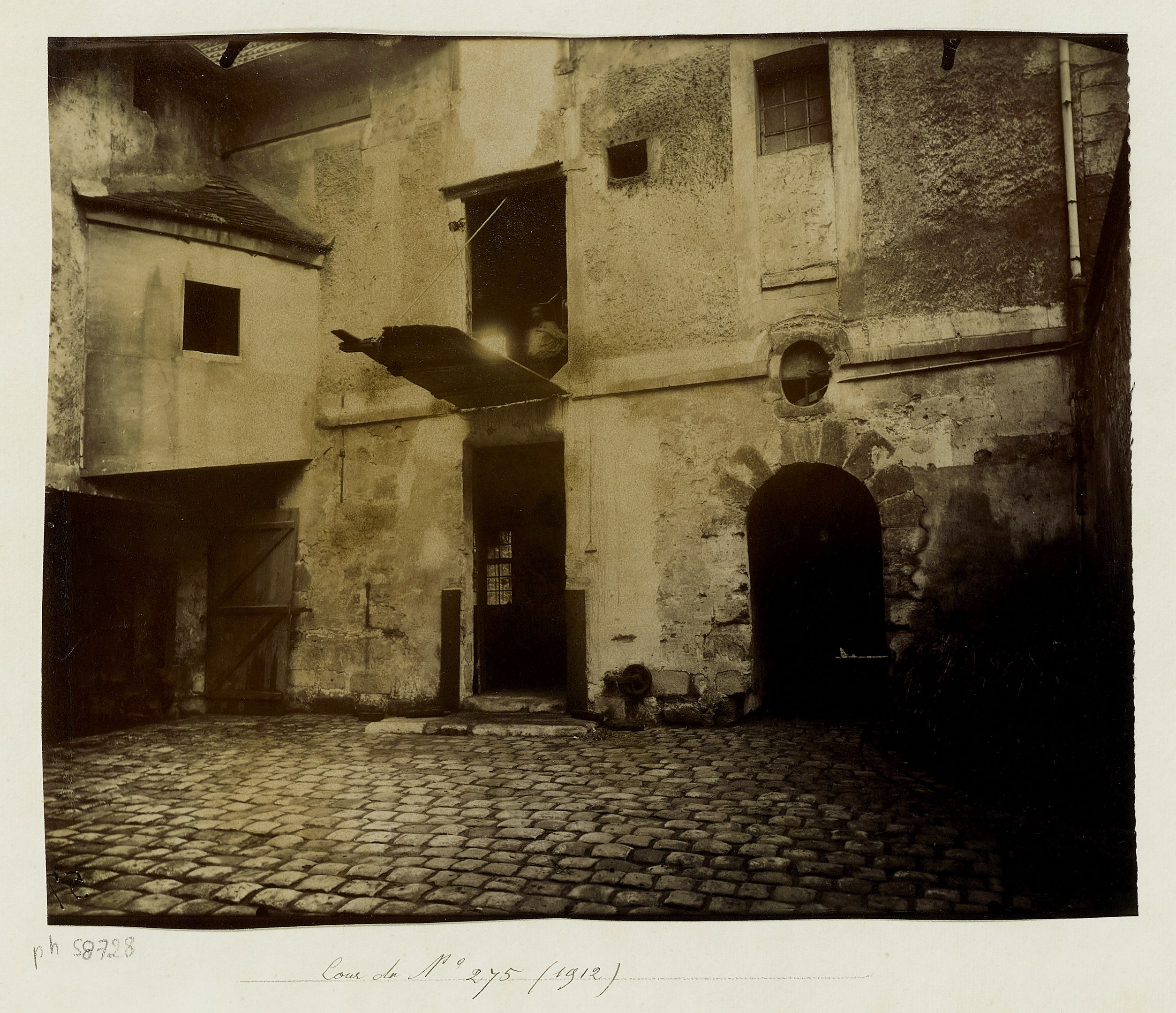 Altes Gebäude mit Fenstern, Türen, einer Rohrleitung und einer Kopfsteinpflasterstraße, mit Text unten.