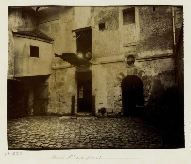 Altes Gebäude mit Fenstern, Türen, einer Rohrleitung und einer Kopfsteinpflasterstraße, mit Text unten.