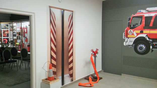 Raum mit einer Wandmalerei eines Feuerwehrautos, einer Tür, einem Verkehrskegel, einem Ständer, einem Teppichboden, Stühlen, Tischen und Feuerlöschern.