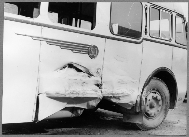 Ein Schwarz-Weiß-Foto eines beschädigten Busses am Straßenrand, dessen Vorderseite eingedellt ist, Fenster zerbrochen und die Fahrertür offen.