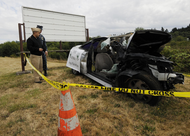 Ein Polizist in Uniform steht neben einem schwer beschädigten Auto mit eingedellter Motorhaube und zersplitterten Fenstern am Straßenrand, mit einem Verkehrskegel und Band in der Nähe, Bäumen und einem bewölkten Himmel im Hintergrund.