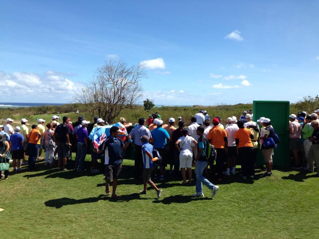 Eine Gruppe von Menschen, einige mit Mützen und Taschen, die auf einem grünen Feld mit Bäumen, Pflanzen, einer bewölktem Himmel und einem Gewässer im Hintergrund stehen, wahrscheinlich bei einem Golfturnier.