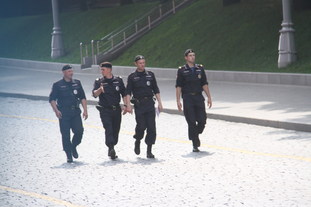 Drei Polizisten in Uniform und Mützen gehen eine Straße mit Pfählen, Geländern, Treppen und Gras im Hintergrund entlang.