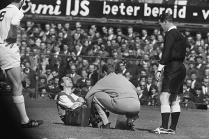 Ein Schwarz-Weiß-Foto eines Schiedsrichters, der mit einem Mann auf dem Feld spricht, umgeben von einer Gruppe von Menschen, mit einem Zaun, einer Fahne und Bäumen im Hintergrund.