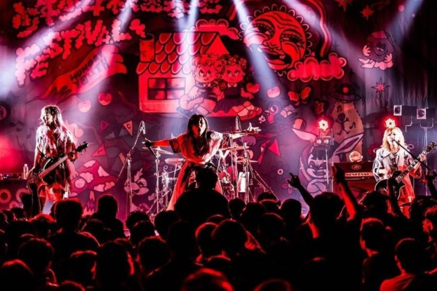 Ein Live-Auftritt der japanischen Rockband Korn auf der Bühne mit Musikern, die Gitarren spielen und in Mikrofone singen, mit einer beleuchteten Wandmalerei im Hintergrund und einer sichtbaren Menge unten.