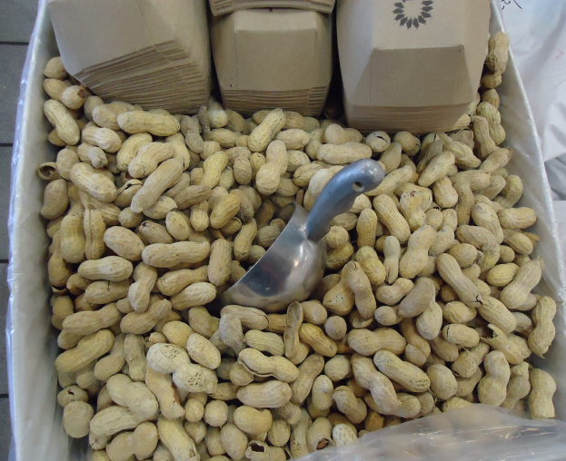 Ein Kasten Erdnüsse auf einem Tisch mit einer Plastikabdeckung und einem Löffel daneben, umgeben von anderen verstreuten Erdnusskisten.