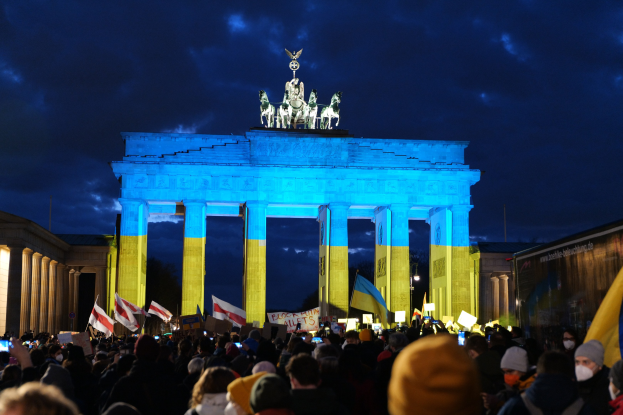 Eine Menge steht vor dem Brandenburger Tor in Berlin, hält Fahnen und Schilder, mit einer Protesttext tragenden Plane auf der rechten Seite.