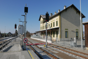 Ein Bahnhofsgebäude mit einem Zug auf den Gleisen, umgeben von Gebäuden, Straßenlaternen, Verkehrspfosten, Verkehrsschildern, Werbetafeln, Zäunen, Bäumen und einem klaren blauen Himmel.