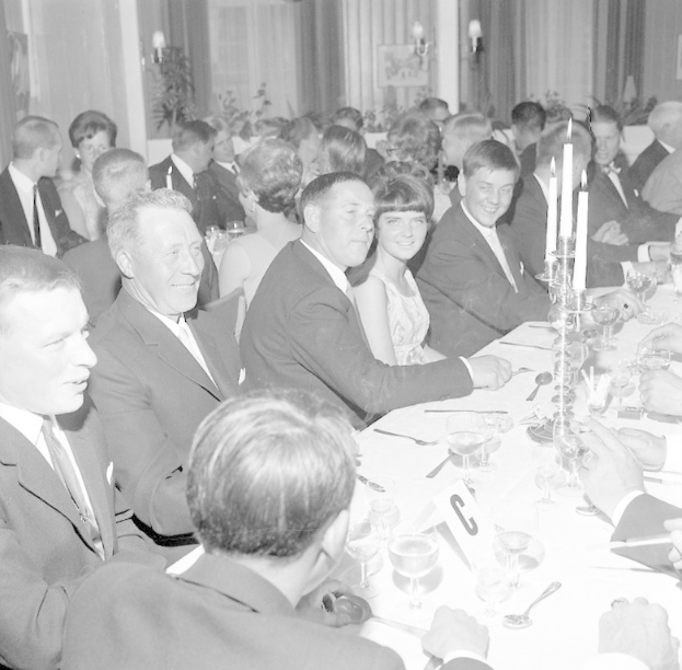 Eine Gruppe von Menschen sitzt um einen langen Tisch mit Essbesteck, beleuchteten Kerzen und warmem Hintergrundlicht.