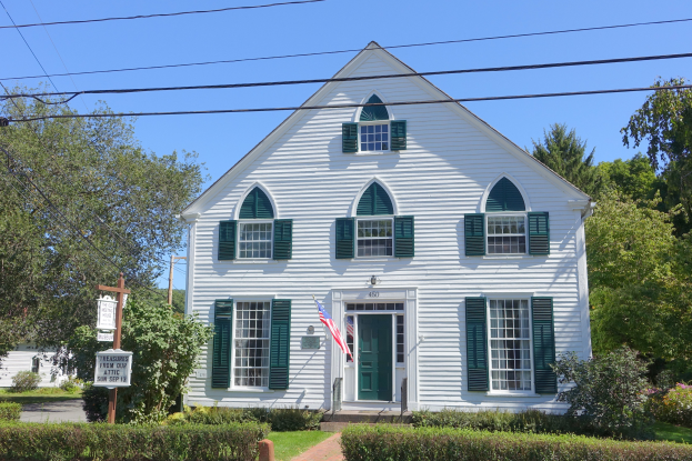 Ein weißes Haus mit grünen Fensterläden und einer Fahne, umgeben von Pflanzen, Gras, einem Weg, Bäumen, einem Schild, Pfählen, Drähten und einem bewölkten Himmel.