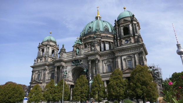 Der Berliner Dom, eine große Kathedrale mit Fenstern, Säulen, Bögen und Statuen, steht prominent in Berlin, Deutschland, mit Menschen in der Nähe, Bäumen, blühenden Pflanzen und einem Turm zu sehen, unter einem bewölkten Himmel.