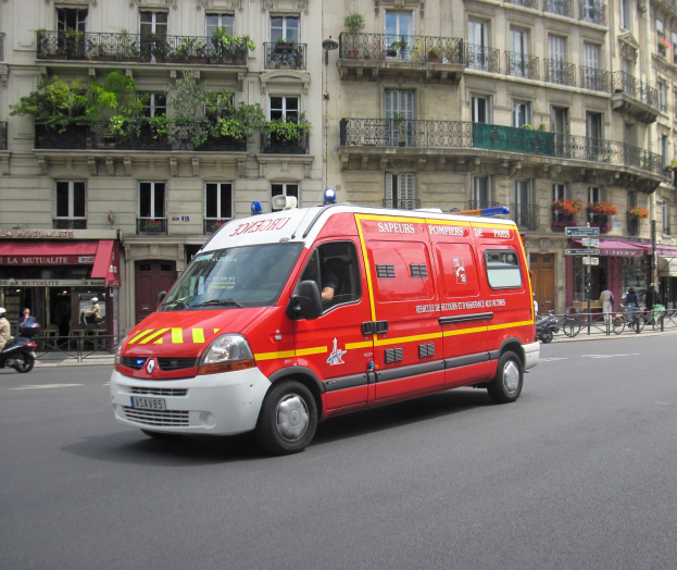 Rettungswagen steht auf der Straße vor einem Gebäude mit Fenstern, Geländern, Pflanzen und anderen Gegenständen, mit ein paar Menschen und Fahrrädern im Hintergrund.