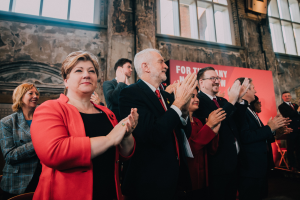Eine Gruppe von Menschen steht vor einer Menge und klatscht begeistert, mit einem Banner mit Text im Hintergrund.