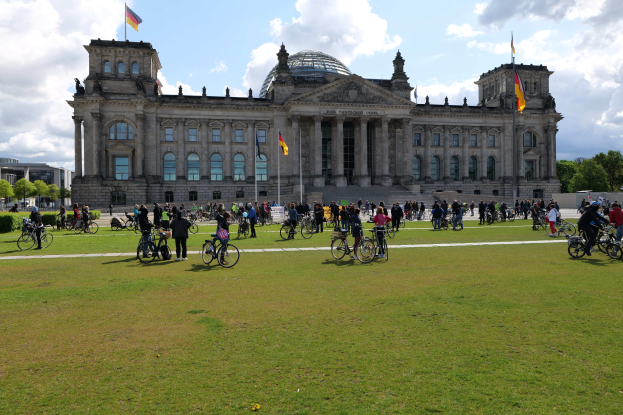 Eine Gruppe von Menschen, die vor dem Reichstaggebäude in Berlin, Deutschland, Fahrräder fährt, mit Bäumen, Pflanzen und einem bewölkten Himmel im Hintergrund.
