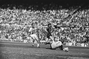 Schwarzes Foto von Männern, die auf einem Feld Fußball spielen, mit Zuschauern im Hintergrund und einem Banner "Bundesliga 1978-79 Ajax 1-1 Hannover 96".