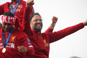 Liverpool-Trainer Jürgen Klopp im roten Mantel feiert mit seinen Spielern nach dem Gewinn der UEFA Champions League, umgeben von zwei medaillentragenden Personen und dem Himmel im Hintergrund.