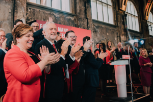 Eine Gruppe von Menschen, wahrscheinlich Liberale, klatscht feierlich vor einer Menge, mit einem Podium, Mikrofon und Texttafel rechts und Stühlen, einer Fahne, Wand, Fenstern und Lichtern im Hintergrund.