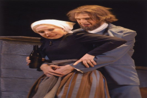 Ein Mann im Anzug mit ernster Miene und eine Frau mit einer Flasche stehen auf einer Bühne mit Text unten und einer Wand im Hintergrund.