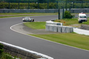 Ein Rennauto fährt eine Rennstrecke entlang mit einem Polizeiwagen im Hintergrund, umgeben von Gras, Pflanzen, Bäumen und Zaun, mit einem Fahrzeug auf der rechten Seite und Text unten.